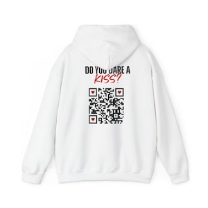 Puede incluir: Sudadera con capucha blanca con el texto "DO YOU DARE A KISS?" en negro y rojo. Debajo del texto hay un código QR con símbolos de corazón. La sudadera tiene capucha y mangas largas.