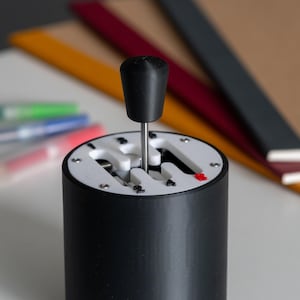 Könnte beinhalten: Ein schwarz-weißer Schalthebelknauf mit zylindrischem Sockel. Die Oberseite des Schalthebelknaufs ist schwarz, und das Schaltschema ist weiß mit einem roten Akzent. Der Hintergrund umfasst Stifte und Notizbücher.