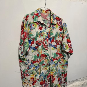 Può includere: Una camicia a maniche corte con un vivace motivo floreale in rosso, verde, blu e giallo su sfondo bianco. La camicia è appesa a una gruccia di metallo contro un muro bianco.