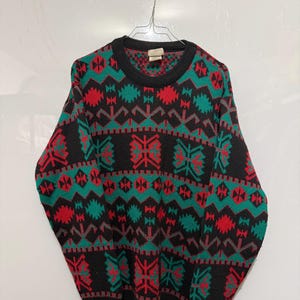 Può includere: Un maglione nero con un motivo geometrico rosso, verde acqua e grigio. Il maglione girocollo è appeso a una gruccia di metallo. Il motivo include forme a diamante e a fiocco di neve.