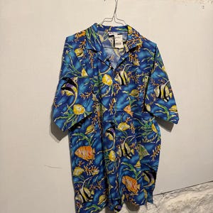 Può includere: Camicia blu a maniche corte con una vivace stampa all-over di pesci tropicali, coralli e alghe. La camicia è appesa a una gruccia di metallo contro un muro bianco. Il design della camicia presenta pesci gialli, arancioni, neri e bianchi.