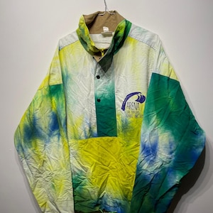 Può includere: Giacca a vento vintage con un motivo tie-dye nelle tonalità del giallo, verde e blu. La giacca presenta un colletto marrone, una chiusura con bottoni e il testo "ARENA SURF MASTERS BARRI-ANGLET". Un cordoncino è visibile in basso.