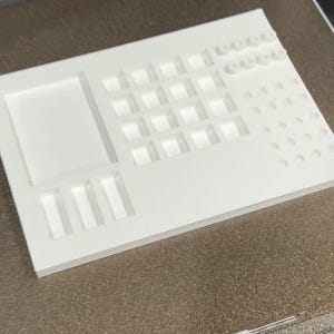 May include: A white, rectangular 3D-printed organizer with various compartments and slots. Features include a large rectangular section, a grid of small squares, and several rows of circular and rectangular holes. The design is clean and modern.