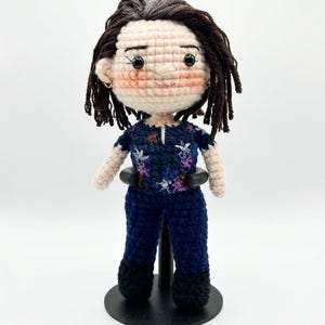 May include: A handmade crochet doll with a light skin tone, dark hair, and a navy blue outfit with floral accents. The doll has a round face, rosy cheeks, and is standing on a black stand. The doll's hair is styled with dark brown dreadlocks.