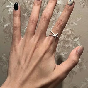 Puede incluir: Una mano con uñas pintadas y un anillo de estrella plateada. Las uñas están pintadas en varios colores, incluyendo negro, marrón y un rosa claro con purpurina. El anillo presenta un diseño de estrella.