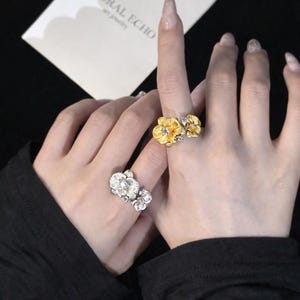 Puede incluir: Primer plano de dos manos que muestran anillos florales. Una mano exhibe un anillo dorado con tres diseños de flores, mientras que la otra mano presenta un anillo plateado con dos diseños de flores. Los anillos están detallados con intrincados patrones de pétalos y acentos brillantes.