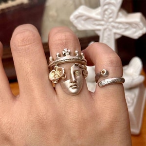 May include: A collection of silver rings. One ring features a crown design, a face, and a flower. Another ring is a wrap-around style with a small yellow stone and the engraved text "ellie m".