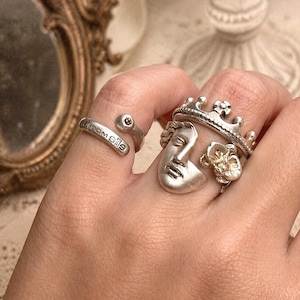 Puede incluir: Una colección de anillos de plata, incluyendo un anillo de corona, un anillo de cara y un anillo con la palabra "bloom oils". Los anillos se llevan en una mano, mostrando sus intrincados diseños y texturas. Los anillos tienen una estética vintage.