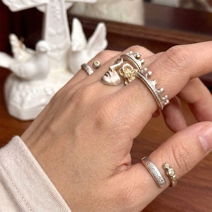 May include: A hand displays several silver rings, including a face design with a crown and a ring with the word "cillemode". The rings have gold accents and small gemstones. A white decorative object is in the background.
