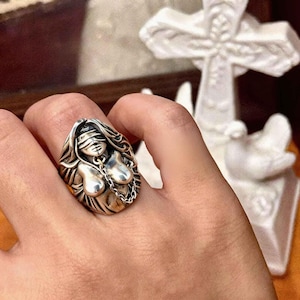 Puede incluir: Un anillo de plata con una mujer con los ojos vendados, pelo largo y una cadena. El anillo tiene un diseño intrincado y se lleva en un dedo. Una pieza de joyería única y distintiva.