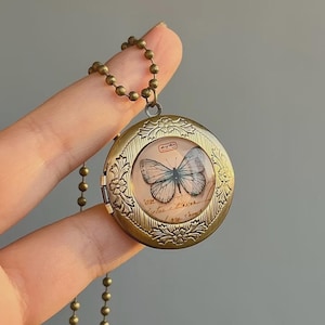 Puede incluir: Un collar con un medallón de latón de estilo vintage, con una ilustración de mariposa. El colgante redondo tiene un diseño floral ornamentado en los bordes y una cadena de bolas. La imagen de la mariposa está sobre un fondo de papel envejecido con escritura manuscrita.
