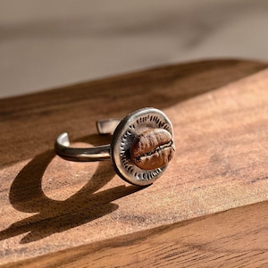 May include: A silver-toned ring featuring a coffee bean set in a circular, textured bezel. The ring's band is open, allowing for adjustable sizing. The coffee bean is a rich brown color, contrasting with the silver metal. The ring is displayed on a wooden surface.