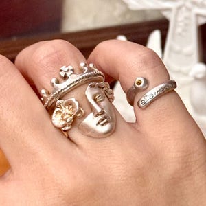 Puede incluir: Una colección de anillos de plata, que incluye un anillo de corona, un anillo con una cara esculpida, un anillo floral y un anillo envolvente con la inscripción "elle moga". Los anillos se llevan en un dedo.