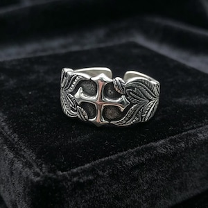 Puede incluir: Un anillo plateado con un diseño abierto. El anillo presenta un diseño de cruz en el centro, rodeado de intrincados patrones de hojas. El anillo se encuentra sobre un fondo oscuro, resaltando sus intrincados detalles.