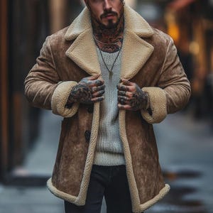 Könnte beinhalten: Eine hellbraune Wildlederjacke mit dickem, cremefarbenem Lammfellfutter, einem breiten Reverskragen und langen Ärmeln mit umgeschlagenen Bündchen. Die Jacke wird über einem hellgrauen Strickpullover getragen und vorne mit dunklen Knöpfen geschlossen.