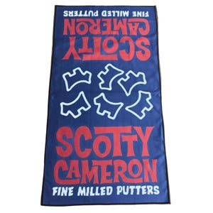 May include: A navy blue golf towel with the words "SCOTTY CAMERON" in red, and "FINE MILLED PUTTERS" in white. The towel features a pattern of white shapes and a black border.