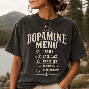 May include: Dark gray t-shirt with the text "DOPAMINE MENU" above a list of outdoor activities. The list includes trails, lake days, campfires, moving water, and no notifications. The shirt has a vintage, worn look.