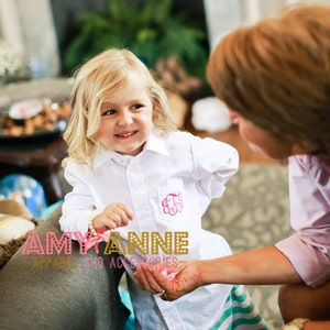 May include: A young girl wearing a white button-down shirt with a pink monogrammed pocket and a green and white striped skirt. She is holding a pink flower in her hand and smiling at the camera. The girl is standing in front of a fireplace with a woman standing behind her.