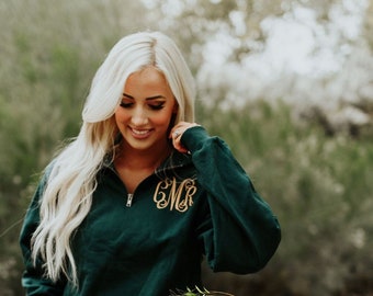 May include: A woman wearing a dark green, quarter-zip sweatshirt with a gold monogram on the chest. She is wearing blue jeans with rips in the knees and brown ankle boots. The sweatshirt has a relaxed fit and is perfect for casual wear.