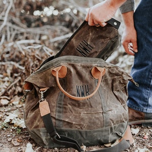 May include: A brown waxed canvas duffel bag with leather handles and accents. The bag has the initials "NWR" embroidered on the front. A matching pouch is being placed inside the bag. The bag is designed for travel.