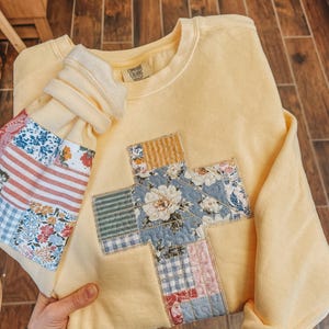 May include: A pale yellow crewneck sweatshirt featuring a large cross applique on the front and matching sleeve patches. The patches are constructed from various quilted fabric squares in floral, striped, and gingham patterns. A Comfort Colors label is visible.