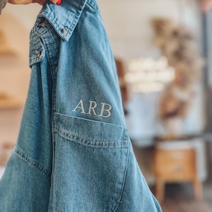 May include: A light blue denim shirt with a pocket and the embroidered letters "ARB" in white. The shirt has a classic collar and button closures. The denim fabric has a slightly faded appearance.