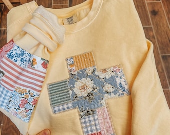May include: A pale yellow crewneck sweatshirt featuring a large cross appliqué on the front and matching sleeve patches. The patches are constructed from various quilted fabric squares in floral, striped, and gingham patterns. A Comfort Colors label is visible.