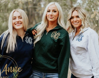 May include: Three women wearing quarter-zip sweatshirts with embroidered monograms. The sweatshirts are navy blue, forest green, and light gray. The women are standing in a wooded area.