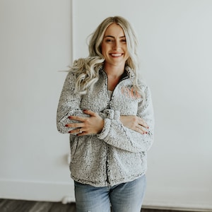 May include: A woman wearing a gray sherpa jacket with a black zipper and an orange monogram. She is smiling and has her arms crossed.