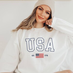 May include: White sweatshirt with a blue "USA" graphic and an American flag with the text "EST. 1776" underneath.