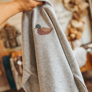 May include: A light gray sweatshirt featuring a small, embroidered cross-stitch design of a mallard duck on the upper chest area. The duck is depicted with a green head, a yellow beak, and a brown body, rendered in a pixelated, stitched style.