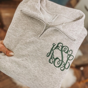 Könnte beinhalten: Ein hellgraues Sweatshirt mit einem Viertel-Reißverschluss-Ausschnitt und einem grünen Monogramm. Das Sweatshirt ist gefaltet und zeigt die gestickten Initialen. Der Stoff hat eine melierte Textur.