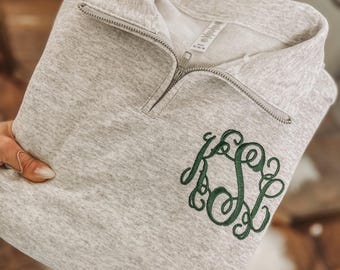 May include: A light gray sweatshirt with a quarter-zip neckline and a green monogram. The sweatshirt is folded, showing the embroidered initials. The fabric has a heathered texture.