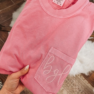 May include: A pink Comfort Colors t-shirt with a pocket featuring embroidered cursive letters "bgl". The shirt is a solid color and has a crew neck. The tag reads "Comfort Colors" and the size is L-G.