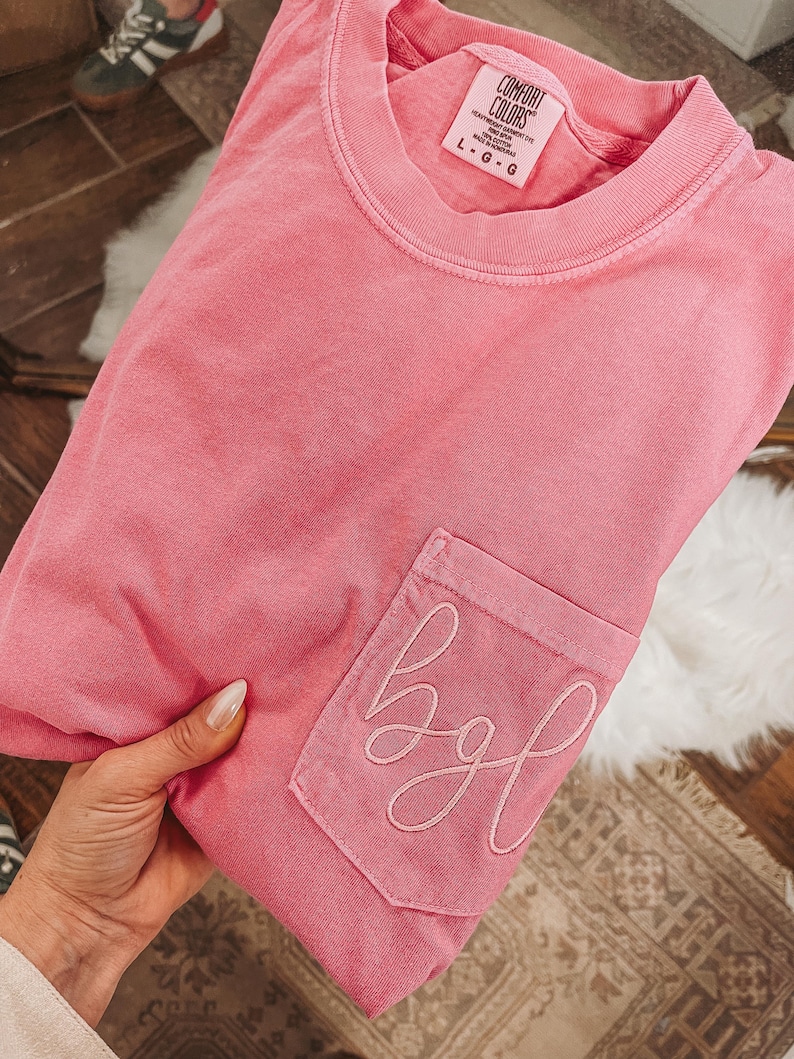 May include: A pink Comfort Colors t-shirt with a pocket featuring embroidered cursive letters "bgl". The shirt is a solid color and has a crew neck. The tag reads "Comfort Colors" and the size is L-G.