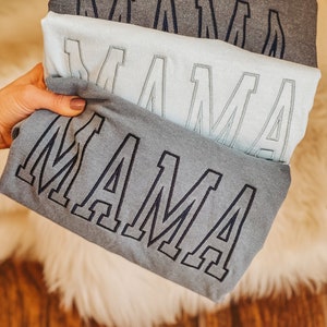 May include: Three grey t-shirts stacked on top of each other. Each shirt has the word "MAMA" embroidered on the front in a large, bold font.