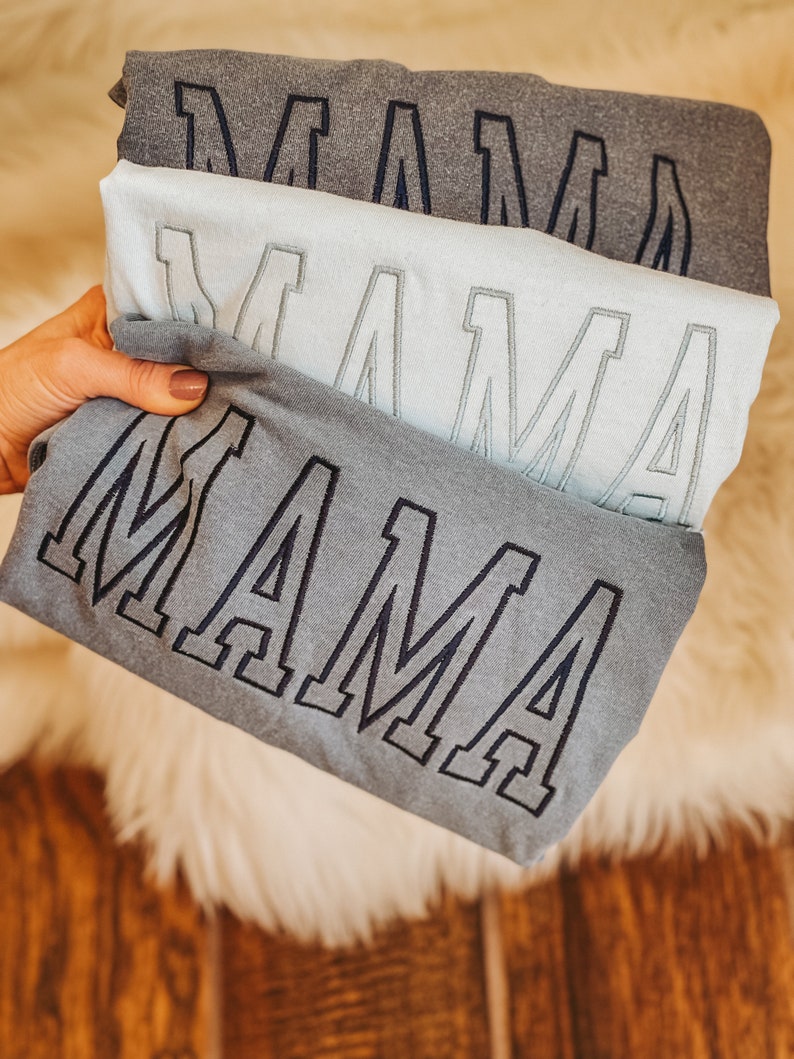 May include: Three grey t-shirts stacked on top of each other. Each shirt has the word "MAMA" embroidered on the front in a large, bold font.