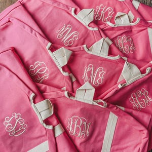 May include: A collection of pink tote bags with white trim and handles. Each bag features a white embroidered monogram. The bags are made of a sturdy material and appear to be folded and stacked.