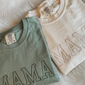 May include: Two t-shirts with the word "MAMA" embroidered on them. The shirts are in different colours, one is green and the other is beige. Both shirts have a tag that says "Comfort Colors" and the size.