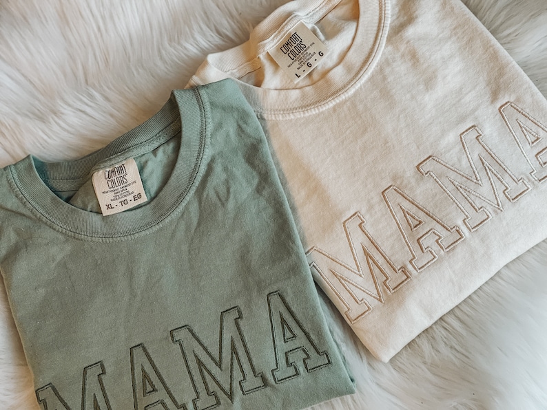 May include: Two t-shirts with the word "MAMA" embroidered on them. The shirts are in different colours, one is green and the other is beige. Both shirts have a tag that says "Comfort Colors" and the size.