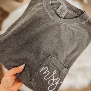 May include: A dark gray t-shirt with a pocket on the front, embroidered with the letters "msg" in white script. The shirt is folded and appears to be made of a soft, comfortable material. The tag reads "2XL - 2TG - 2XG".