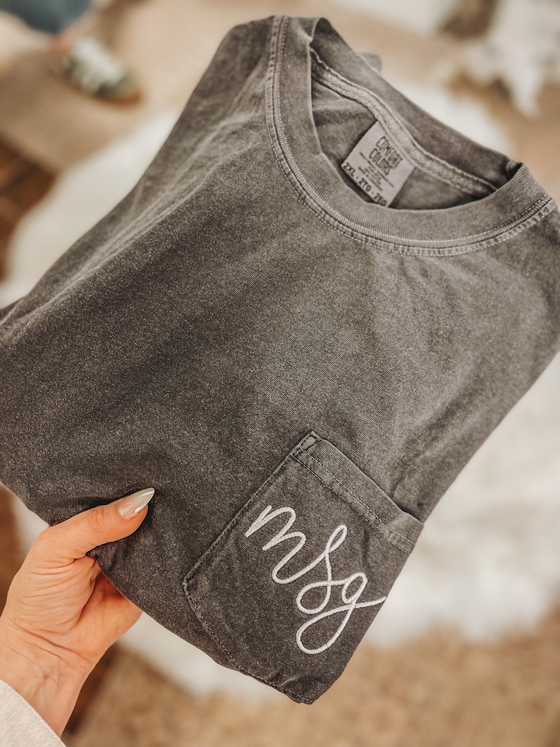 May include: A dark gray t-shirt with a pocket on the front, embroidered with the letters "msg" in white script. The shirt is folded and appears to be made of a soft, comfortable material. The tag reads "2XL - 2TG - 2XG".
