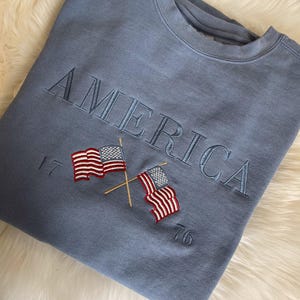 May include: A blue sweatshirt with the word "AMERICA" embroidered across the chest in a matching blue thread. Below the word are two embroidered American flags with red and white stripes and blue stars, and the numbers "17" and "76".