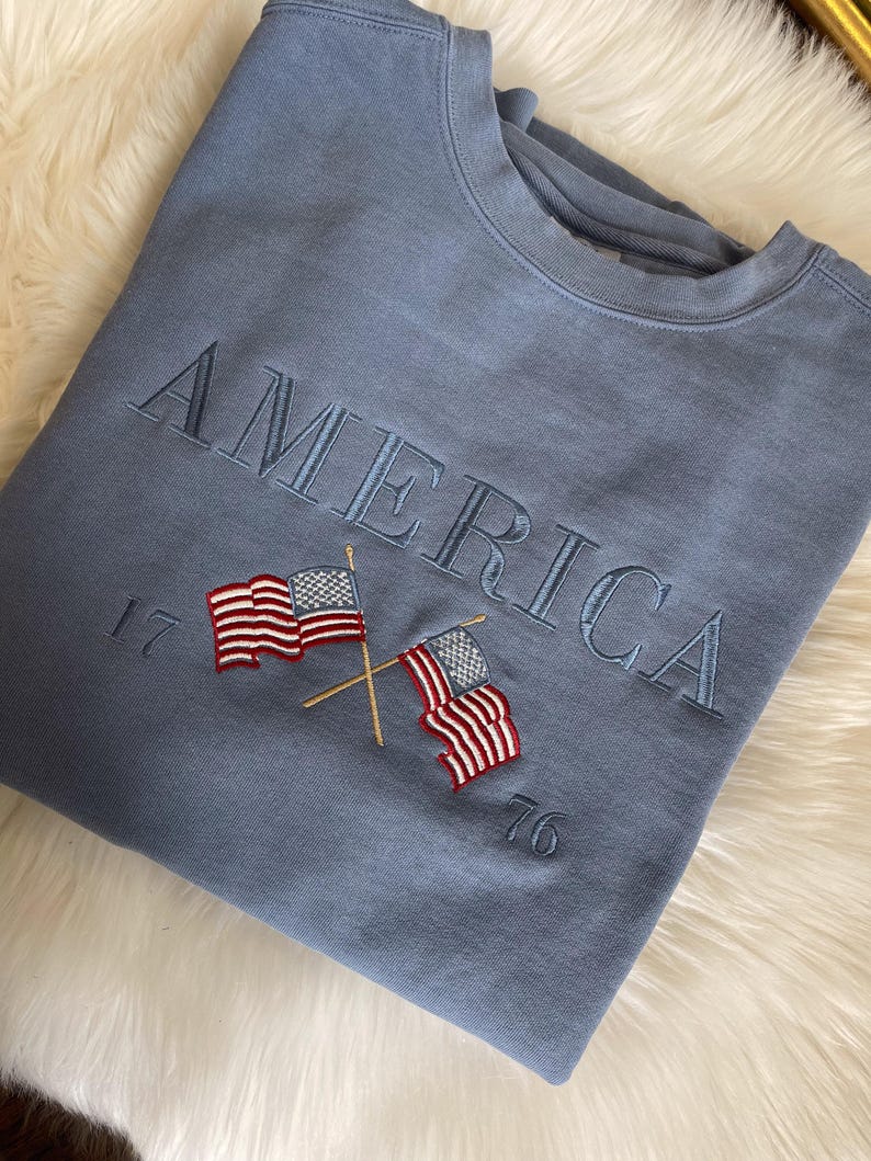 May include: A blue sweatshirt with the word "AMERICA" embroidered across the chest in a matching blue thread. Below the word are two embroidered American flags with red and white stripes and blue stars, and the numbers "17" and "76".