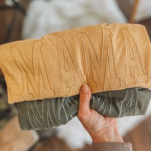 May include: Two folded shirts with the word "MAMA" embroidered on them. The top shirt is a light brown colour and the bottom shirt is a dark green colour.
