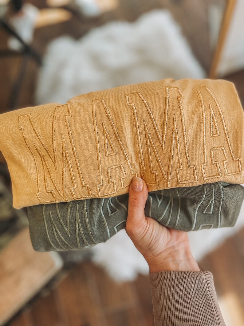 May include: Two folded shirts with the word "MAMA" embroidered on them. The top shirt is a light brown colour and the bottom shirt is a dark green colour.