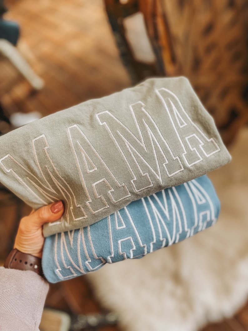 May include: Two sweatshirts with the word "MAMA" embroidered in white on a light blue and a sage green background.