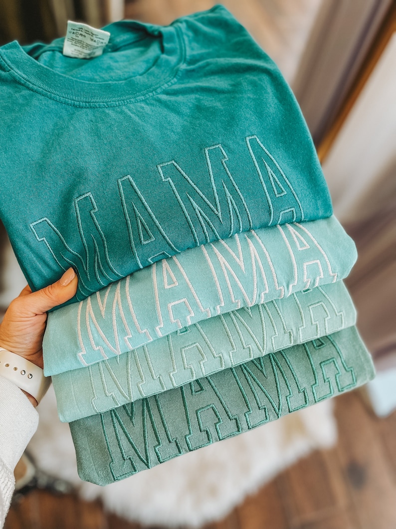 Pode incluir: Pilha de quatro t-shirts azul turquesa com a palavra "MAMA" bordada em branco em cada camiseta.