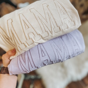 May include: Two sweatshirts stacked on top of each other. The top sweatshirt is cream coloured with the word "MAMA" embroidered in gold. The bottom sweatshirt is lavender with the word "MAMA" embroidered in a darker shade of lavender.