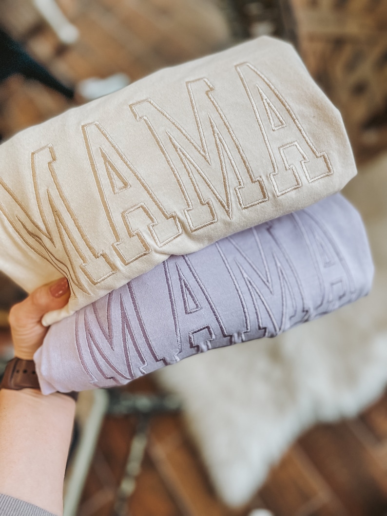 May include: Two sweatshirts stacked on top of each other. The top sweatshirt is cream coloured with the word "MAMA" embroidered in gold. The bottom sweatshirt is lavender with the word "MAMA" embroidered in a darker shade of lavender.